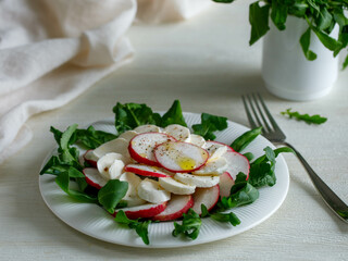 Plate with healthy spring vegetarian salad with radish, mozzarella cheese and rucola with olive oil. top view on white wooden background with napkin, summer food