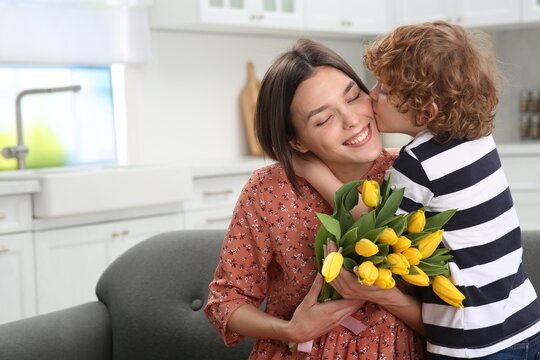 Little son kissing and congratulating his mom with Mother`s day at home, space for text. Woman holding bouquet of yellow tulips