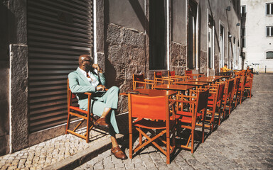 Naklejka premium A stylish Black man bald-headed sits alone, lost in thought, on a closed terrace in Lisbon, Portugal; the outdoor furniture remains, shining in the bright sunshine against the traditional pavement