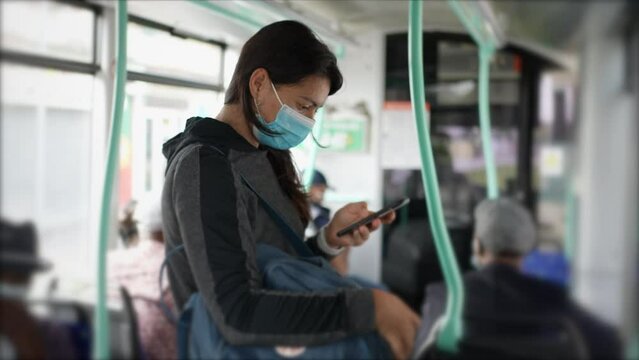 Woman standing inside bus wearing surgical face mask while checking smartphone device inside public transportation during commute