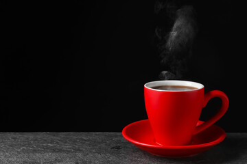 Red cup with hot steaming coffee on grey table against black background, space for text