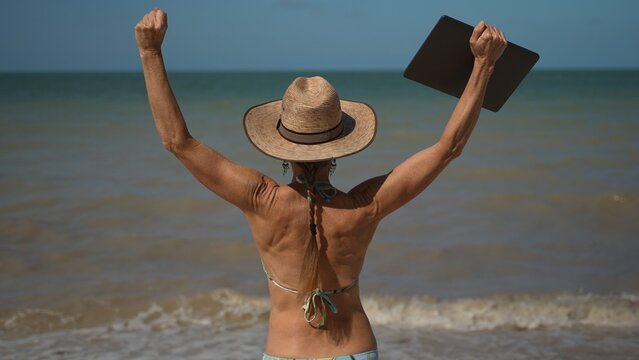 Pretty mature senior woman in hat sunglasses and bikini holding laptop at the beach showing freedom of being a digital nomad in tropical place.