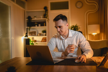 Fototapeta premium A young man is working late or studying at his home office while drinking coffee and using his mobile phone