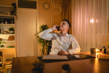 A young man is working late or studying at his home office while drinking coffee and using his mobile phone