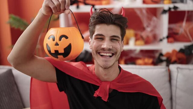 Young hispanic man wearing devil costume holding halloween pumpkin basket at home