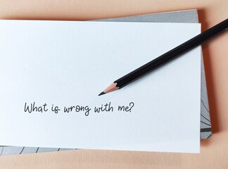 Pencil on note paper with text written WHAT IS WRONG WITH ME?, concept of moment of self doubt, feeling stuck or overwhelmed circumstances of questioning whether one can make it through.