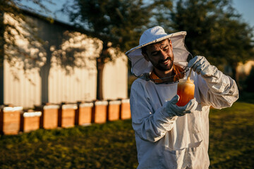 A beekeeper man in his 30s is holding a glass of high-quality bee honey in her hands, wearing...