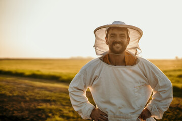 Portrait of a handsome beekeeper in a protective uniform standing outdoors in the field. Copy space - obrazy, fototapety, plakaty