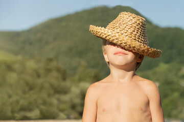 Portrait of a smiling boy. He pulled a straw hat over his eyes. The bank of the river, on the background of the mountains.