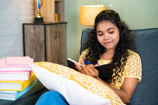 Smiling indian young girl writing personal diary while sitting at home before sleeping - concept of hobbies, imagination and routine