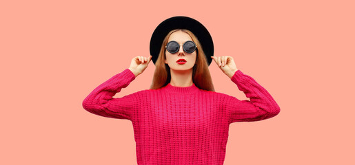 Portrait of stylish young woman model blowing her red lips sends kiss wearing pink knitted sweater and black round hat isolated on background