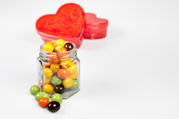 A red heart-shaped translucent gift box and a glass jar with colorful chocolate balls. Confess your love with all your heart