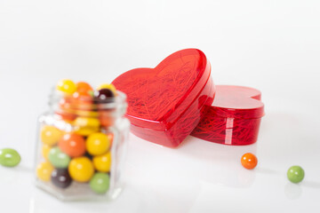 A red heart-shaped translucent gift box and a glass jar with colorful chocolate balls. Confess your love with all your heart