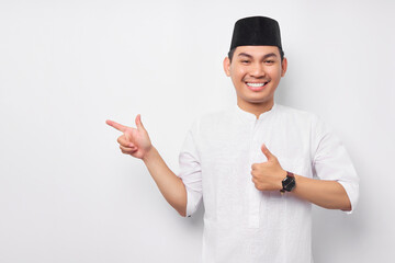 Happy smiling handsome young Asian Muslim man pointing finger aside empty space and giving thumbs up sign gesture isolated over white background. People religious Islamic lifestyle concept