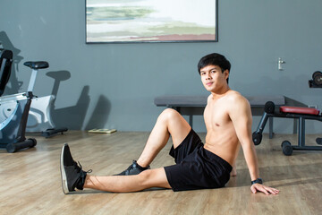 Asian handsome young man is warming up his body by stretching his legs while looking the camera in fitness center.