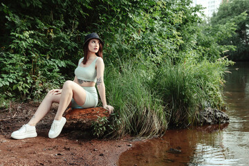 Young jewish woman in clothes and black hat sitting on stone near river on nature, looking away....