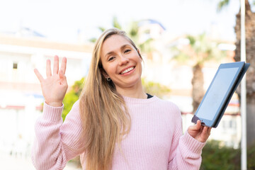 Young blonde woman holding a tablet at outdoors saluting with hand with happy expression