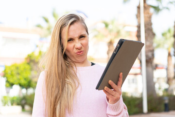 Young blonde woman holding a tablet at outdoors with sad expression