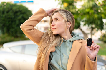 Young blonde woman holding car keys at outdoors having doubts and with confuse face expression