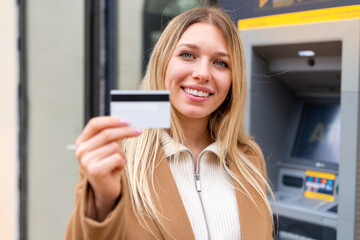 Young pretty blonde woman holding a credit card at outdoors with happy expression
