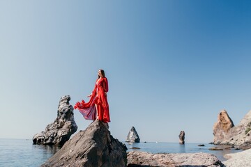 Woman travel sea. Young Happy woman in a long red dress posing on a beach near the sea on background of volcanic rocks, like in Iceland, sharing travel adventure journey