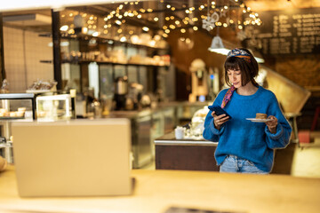 Young stylish woman using phone while carrying her order at modern coffee shop. Concept of modern lifestyle and digital work at cafe