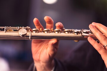A close up portrait of the fingers of a hand of a flutist musician gripping down on the valves of a metal silver flute to play a note of a musical piece during a concert.
