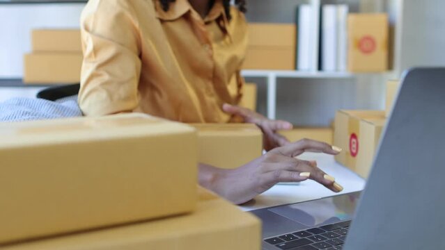 African young woman owner of a start up business designing and selling clothes online checking orders on laptop and preparing for delivery.