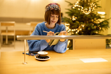 Young adult woman works on a digital tablet while sitting at modern coffee shop in yellow tones with Christmas tree on background. Concept of remote creative work online