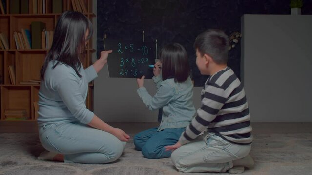 Clever cute elementary age Asian girl writing math division solution on portable chalkboard, while caring charming mother and preadolescent children studying mathematics during homeschooling.