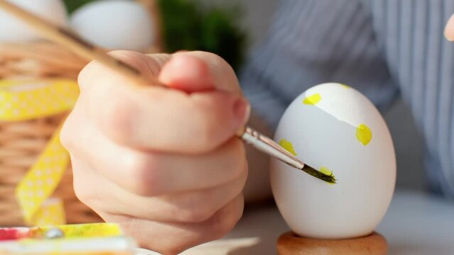 Happy Easter concept. Young womans hand draws a yellow dots pattern on a chicken egg with a brush and paints. Decoration for the spring holiday. Preparation for the celebration.