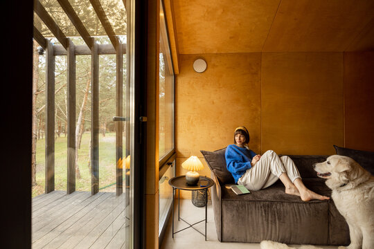 Woman reads a book while sitting relaxed on a couch by the window with her dog at wooden cottage in forest. Concept of home comfort and rest in a house on nature. Wide angle view
