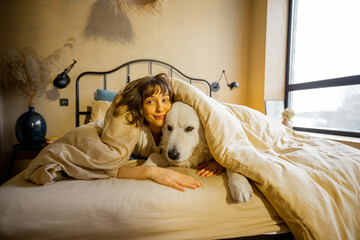 Young woman hugs with her white adorable dog while lying covered with blanket in bed at home. Concept of friendship with pets and home coziness