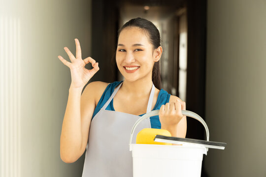 Happy smiling Asian cleaning service worker woman giving ok hand gesture, concept image of domestic helper holding cleaning supplies, ready for work in good working condition