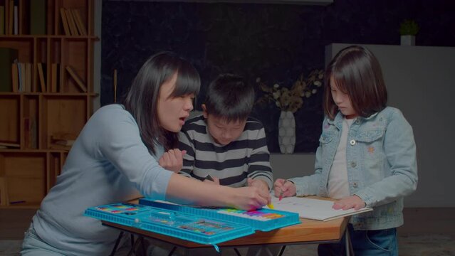 Positive attractive Asian mother and adorable preadolescent children developing creative skills and artistic imagination, designing picture on paper sheet with colorful felt tip pens in domestic room.