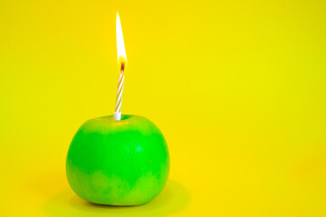 A candle burns in a green apple instead of a cake on a yellow background. 