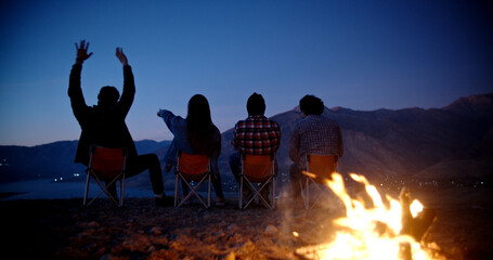 Silhouettes of four friends sitting in chairs near bonfire on top of mountains, watching the sun set and chatting, enjoying their trip - friendship, youth concept 