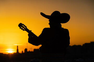 Silhouette of a mexican musician mariachi with a guitar on a background of city panorama.