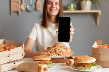 Indoor shot of pretty positive woman with brown hair wearing white T-shirt sitting at table with...
