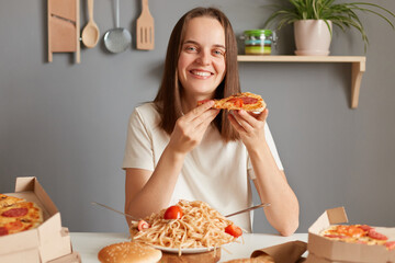 Indoor shot of happy girl eating a pizza, holding a pizza in her hand and staring at camera with satisfaction, having dinner, enjoying delicious unhealthy dish.