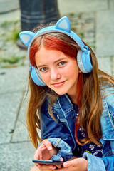 A cute, beautiful teenage girl in blue headphones with cat ears listens to music on a sunny warm day