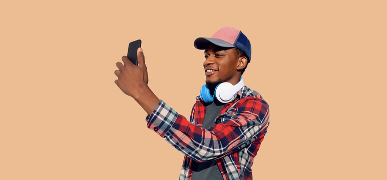 Portrait of modern smiling young african man taking selfie with smartphone listening to music in headphones wearing baseball cap isolated on brown background