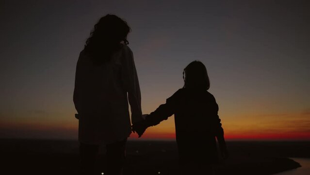 Silhouette of mother and daughter on the background of the sky in the evening after sunset, mother takes her daughter's hand watching the sunset, family love. ProRes