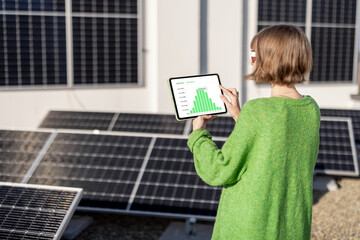 Young woman monitors energy production from the solar power plant with a digital tablet. View on tablet screen with running program. Concept of new technologies in alternative energy - obrazy, fototapety, plakaty