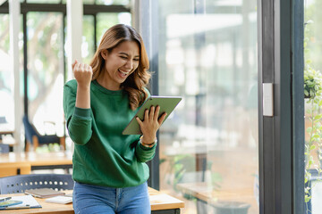 Excited beautiful asian woman looking at digital tablet, young woman with touchpad Excited happy raised arms to celebrate business success - obrazy, fototapety, plakaty