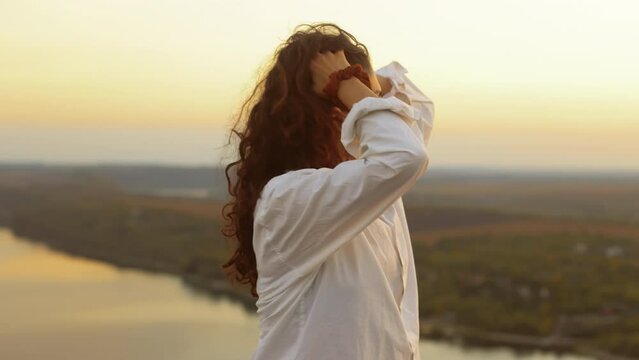 Portrait of a young pretty smiling woman standing in nature against a beautiful view at sunset in the evening, the girl straightens her curly dark hair.