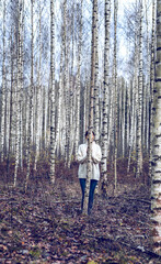 Woman dressed in white knitted pullover and jeans is standing behind thin birch tree in the forest at late autumn