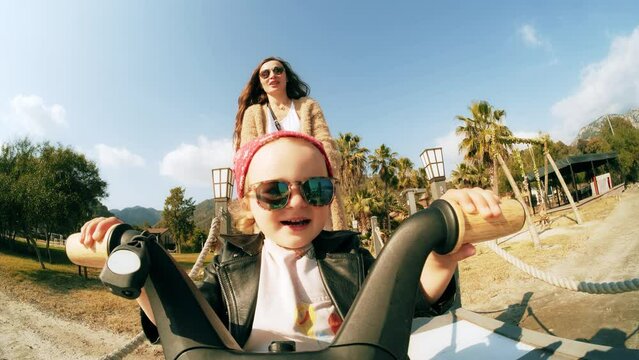 Happy little baby in biker outfit rides her tricycle guided by her mom