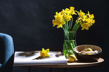 interior, holidays and home decor concept - close up of modern blue chair, easter eggs in bowl, daffodil flowers and magazine on table in dark room