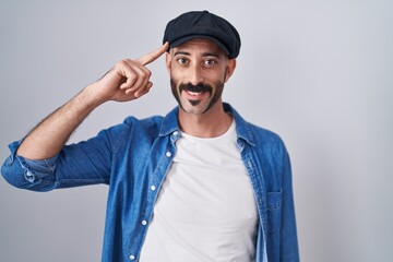 Hispanic man with beard standing over isolated background smiling pointing to head with one finger,...